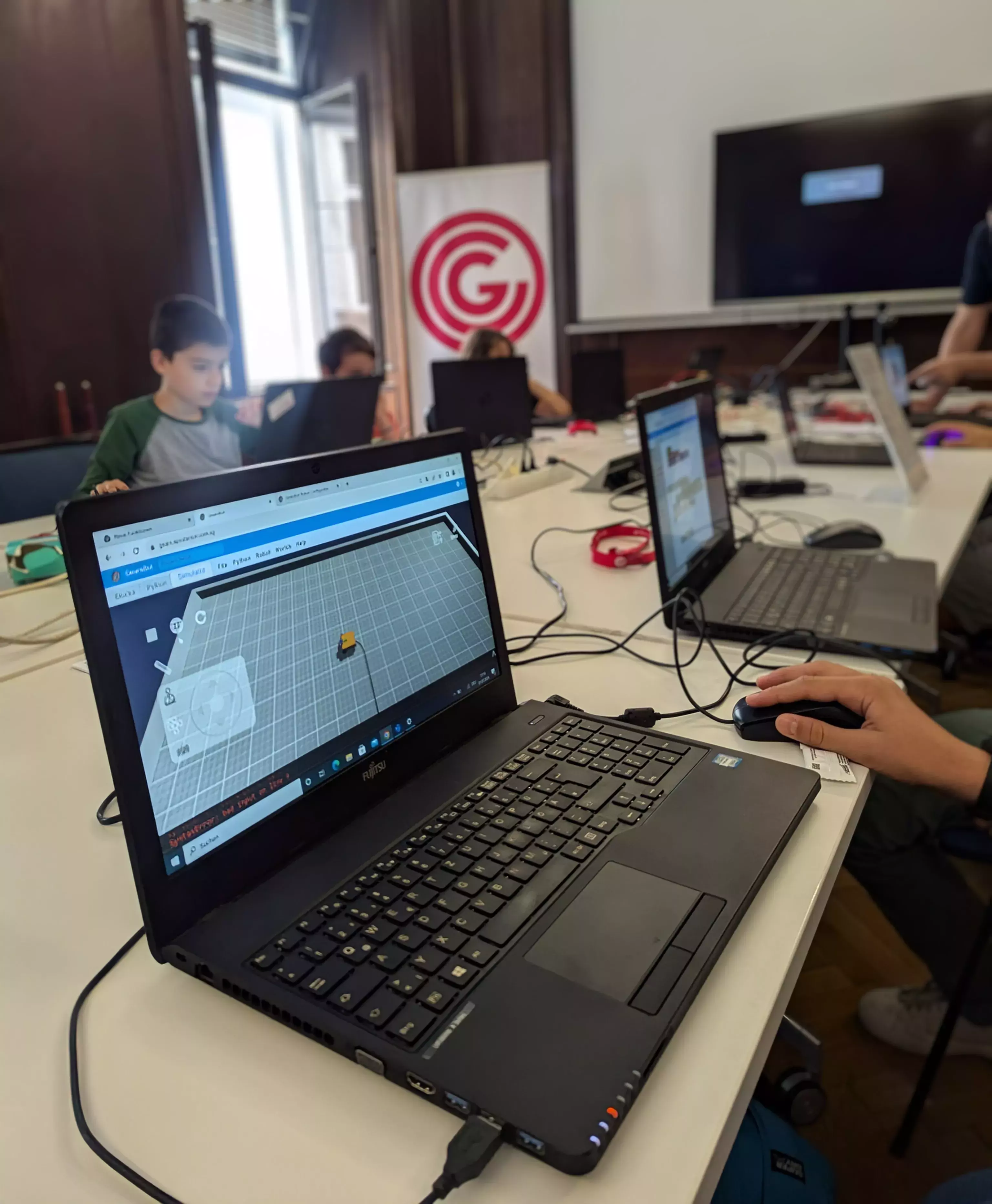 Laptop zu sehen: GearsBot Simulation. Im Hintergrund weitere Laptops und Kinder sowie OCG Banner.