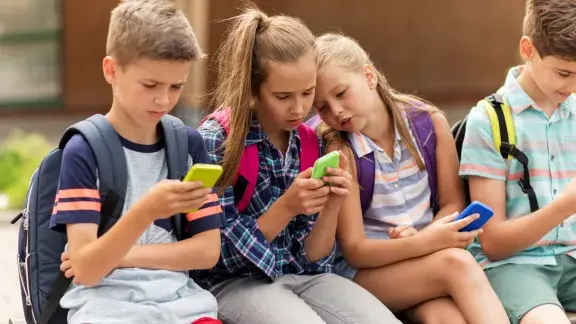 Vier Schüler*innen (ca. 10-jährig), 2 Buben, 2 Mädchen sitzen nebeneinander und schauen auf ihre Smartphones, alle tragen Schulrucksäcke